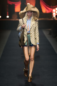Model walking a fashion runway in a large beige hat, patterned blazer, blue shirt, green fringed shorts, and brown buckled boots with a fringed bag.