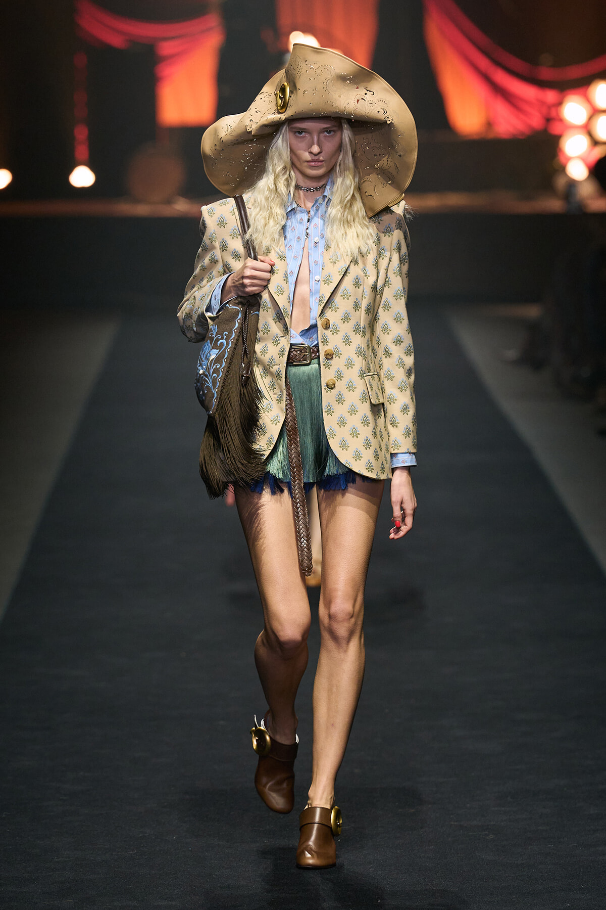 Model walking a fashion runway in a large beige hat, patterned blazer, blue shirt, green fringed shorts, and brown buckled boots with a fringed bag.