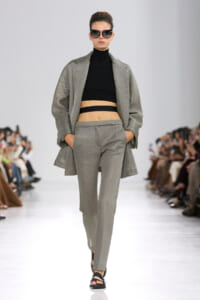 Runway model in a light gray suit with a black cropped top, oversized blazer, and dark sunglasses walking the catwalk.