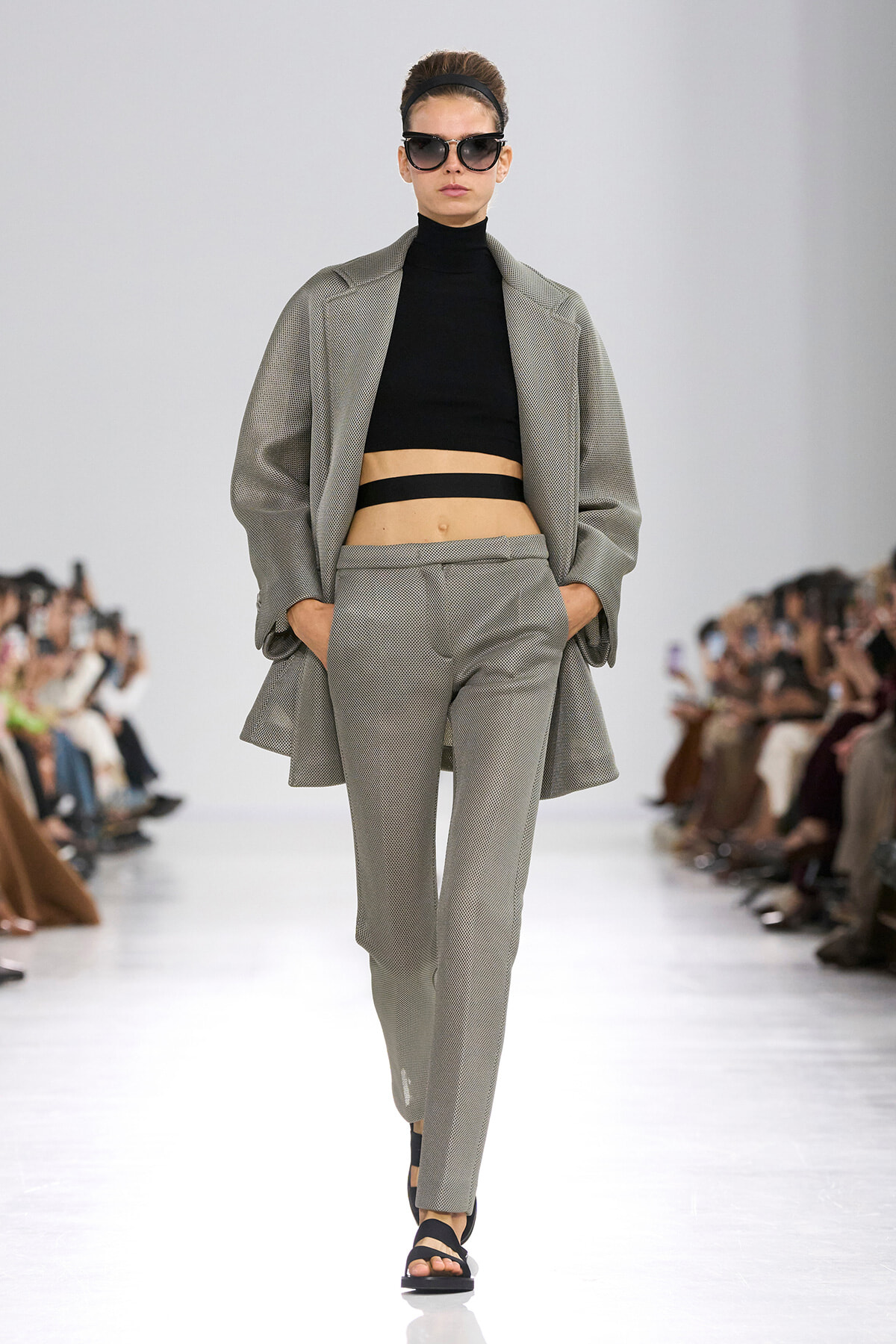 Runway model in a light gray suit with a black cropped top, oversized blazer, and dark sunglasses walking the catwalk.