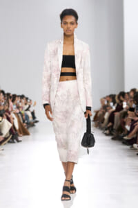 Model walks a runway in a pale pink floral suit with a black bandeau top and a small black handbag.