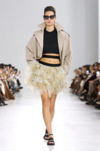 Model walking a runway in a beige oversized trench, black cropped sweater, and a voluminous pastel floral tulle skirt with a black belt and sandals, plus dark sunglasses.