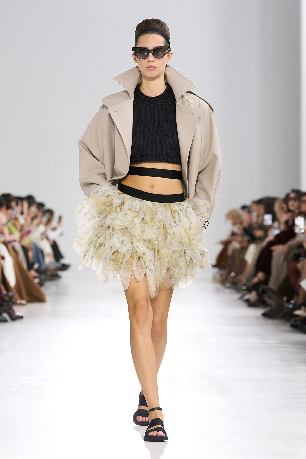 Model walking a runway in a beige oversized trench, black cropped sweater, and a voluminous pastel floral tulle skirt with a black belt and sandals, plus dark sunglasses.