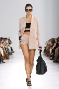 Model on a runway wearing a pale pink blazer and matching shorts with a black bandeau top, oversized sunglasses, and a large black bag.