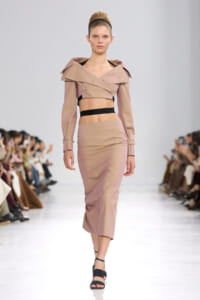 Model walks the runway in a dusty pink cropped top with an oversized collar and a matching high-waisted skirt, black waistband visible at midriff.