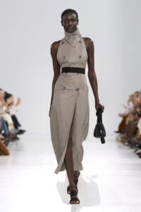 Model walks the runway in a beige sleeveless trench dress with a high collar, black belt, and wrap skirt with a front slit, holding a small black handbag.