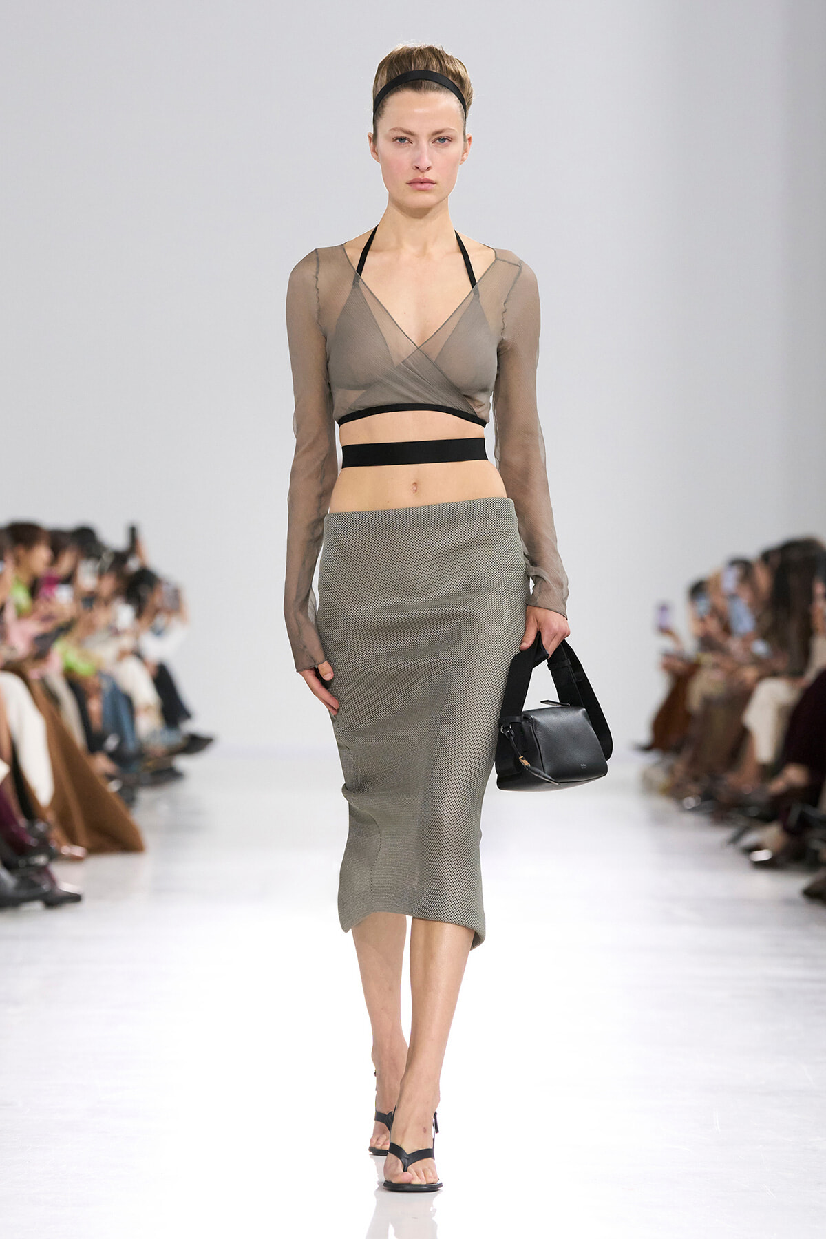 Model walking the runway in a sheer taupe wrap-top with black bands and a gray pencil midi skirt, carrying a black handbag.