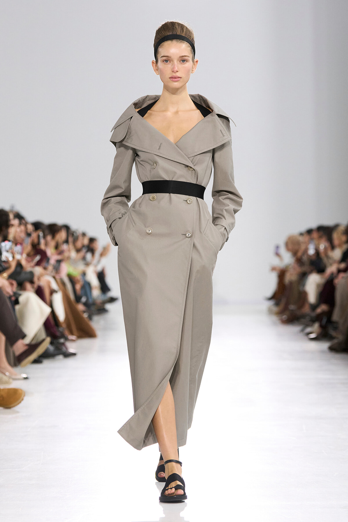Model walking a runway in a beige trench coat with a black belt, double-breasted, during a fashion show.