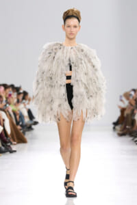 Model walks the runway in a voluminous white feathered cape over a black bodysuit, with a black headband.