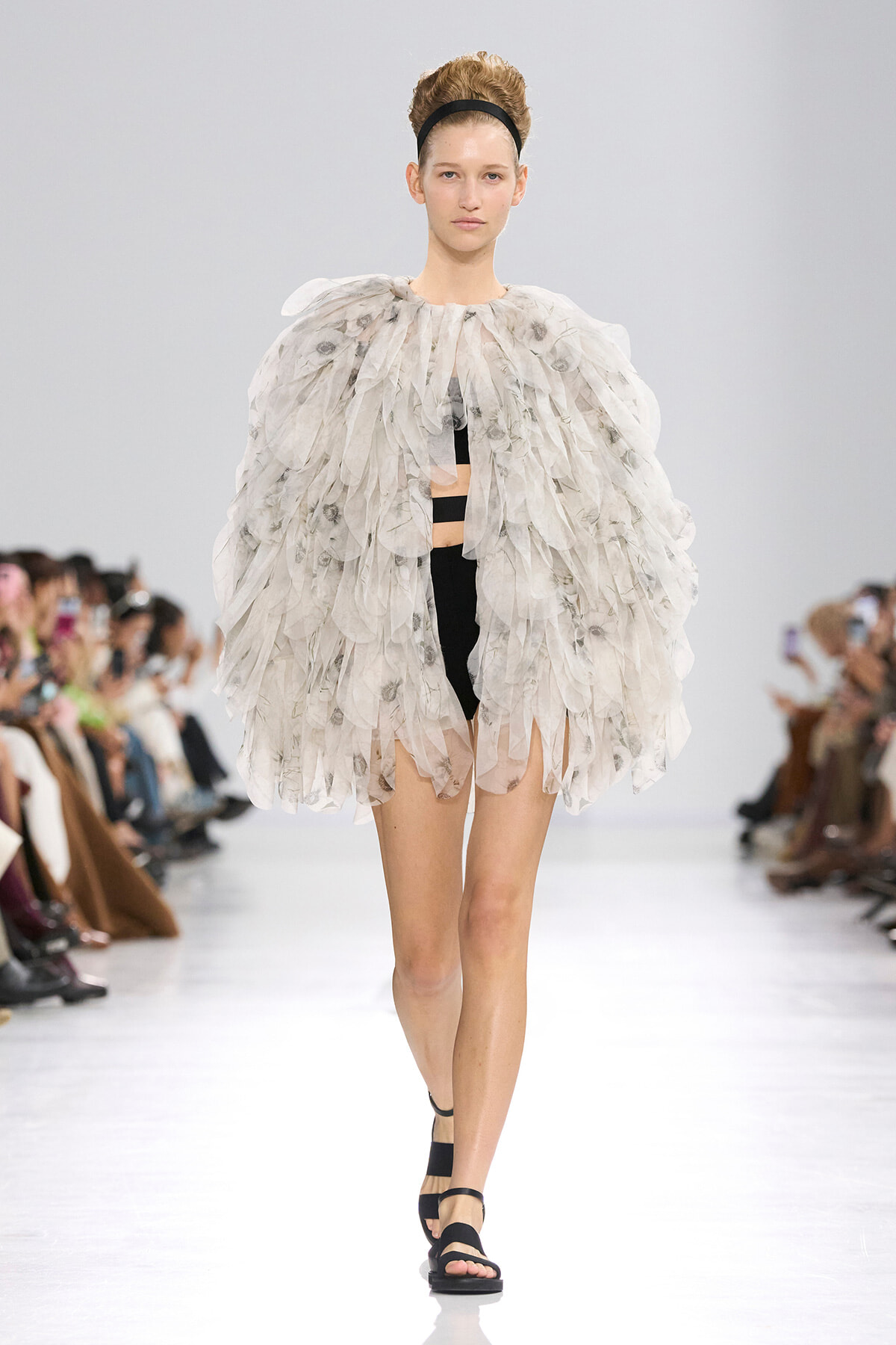 Model walks the runway in a voluminous white feathered cape over a black bodysuit, with a black headband.