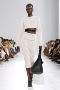 Model walking a runway in a cream knit cropped sweater and matching knit skirt, with a wide black waistband showing at the midriff, carrying a large black handbag and wearing black sandals.