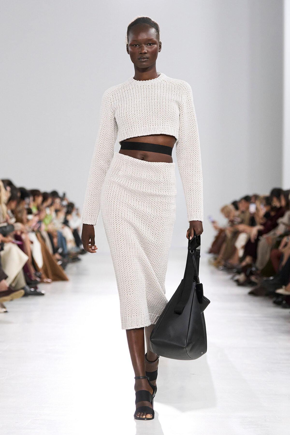 Model walking a runway in a cream knit cropped sweater and matching knit skirt, with a wide black waistband showing at the midriff, carrying a large black handbag and wearing black sandals.