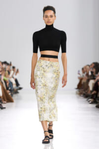 Model walks a fashion show in a black cropped turtleneck and a pale yellow floral midi skirt, black sandals, on a white runway with seated audience.
