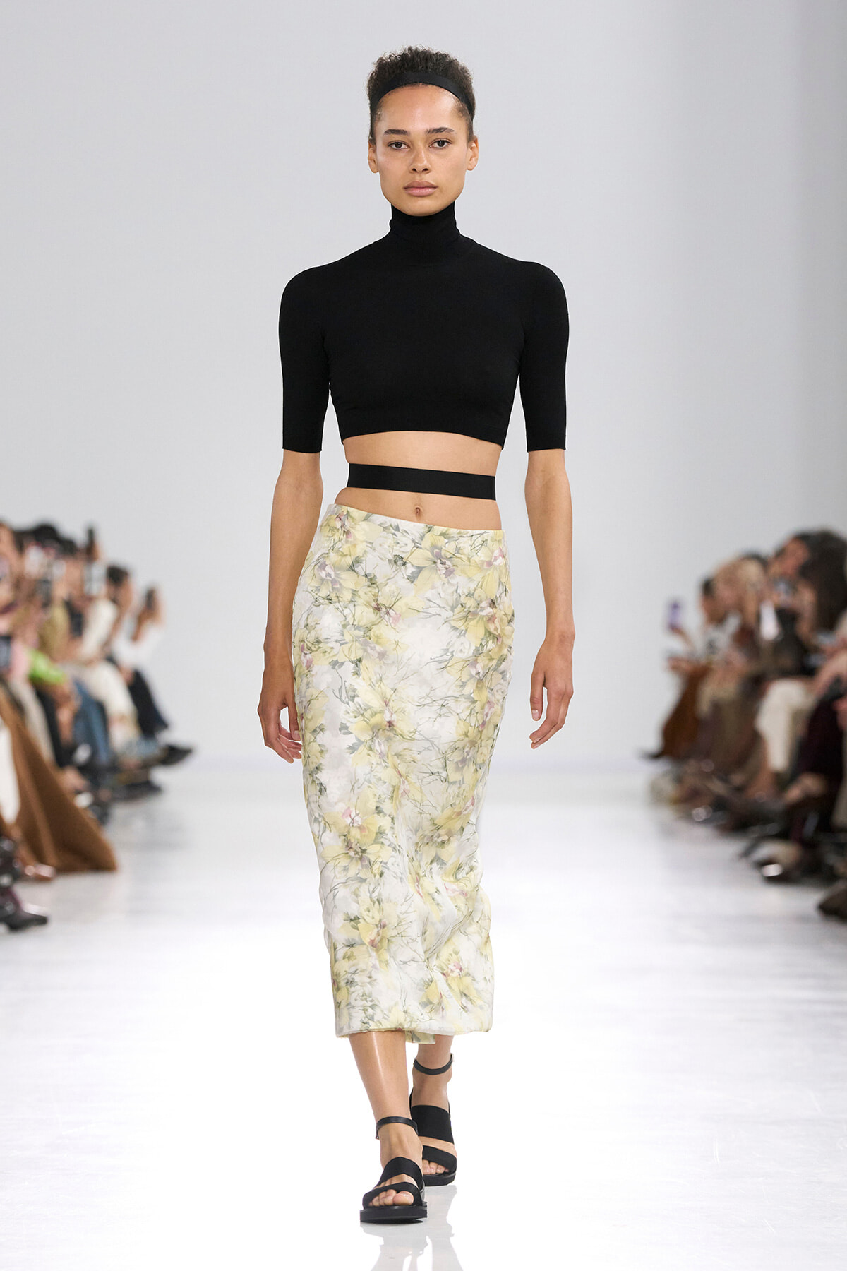 Model walks a fashion show in a black cropped turtleneck and a pale yellow floral midi skirt, black sandals, on a white runway with seated audience.