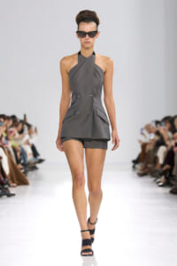 Model walking a runway in a gray halter-neck, double-breasted romper with matching shorts and black sandals, wearing large sunglasses.
