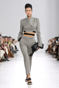 Model walks the runway in a gray cropped blazer and matching high-waisted pants with black waistbands, carrying a black clutch.