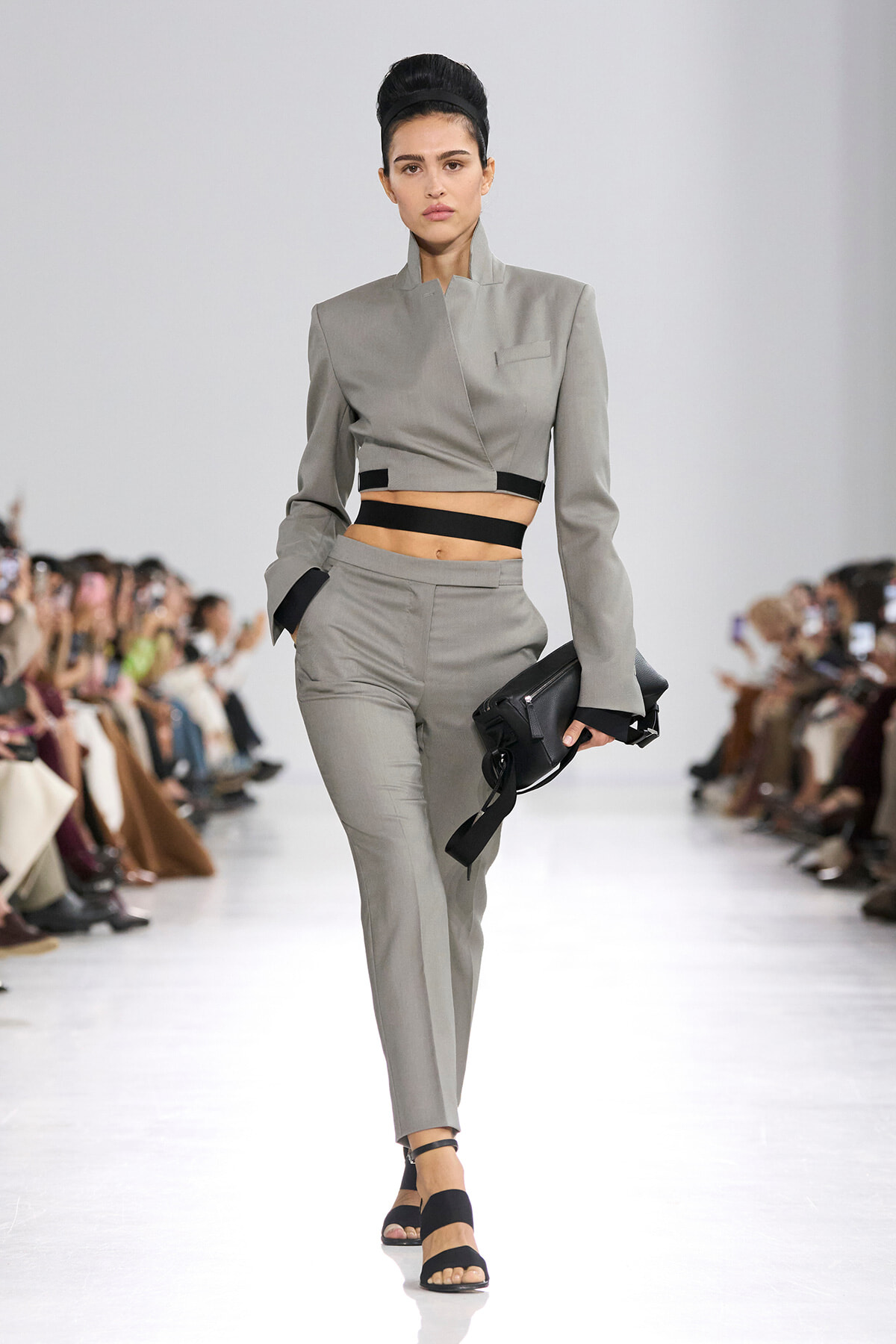 Model walks the runway in a gray cropped blazer and matching high-waisted pants with black waistbands, carrying a black clutch.