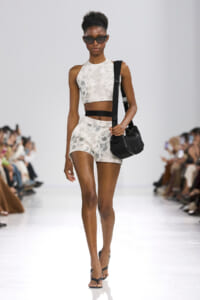 Runway model wearing a pale floral crop top and matching shorts, with a black crossbody bag and dark sunglasses.
