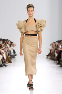 Model walks the runway in a beige two-piece dress with oversized puff sleeves and a black waist belt, black sandals.