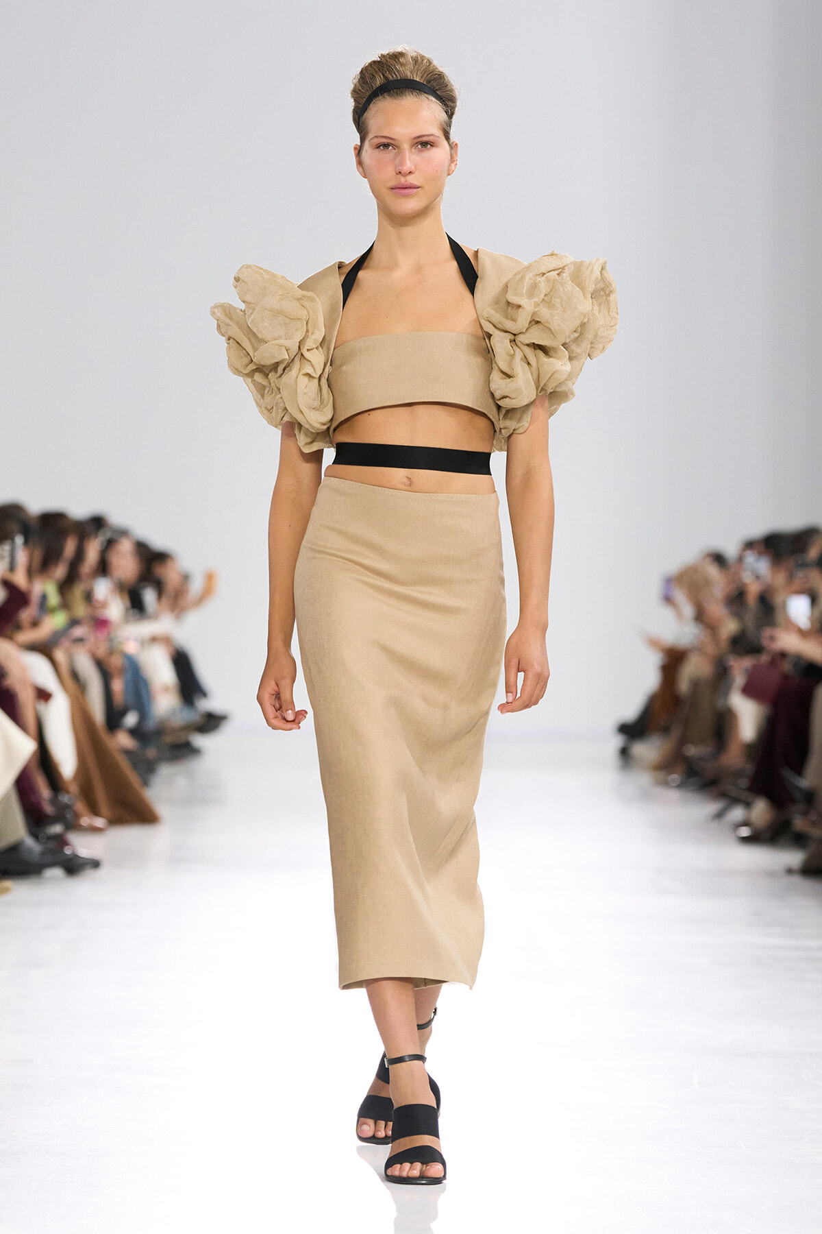 Model walks the runway in a beige two-piece dress with oversized puff sleeves and a black waist belt, black sandals.