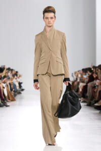 Model walks a fashion runway wearing a beige tailored pantsuit (single-button blazer with peplum-like pockets and wide-leg trousers), paired with a black headband and a large black handbag.