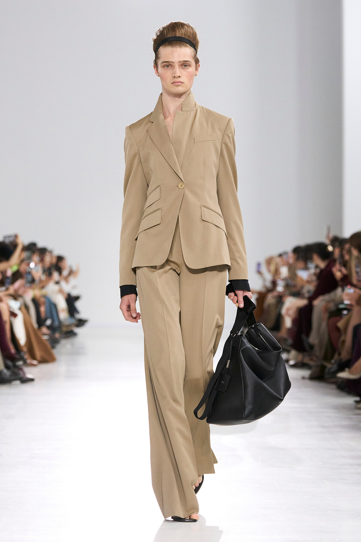 Model walks a fashion runway wearing a beige tailored pantsuit (single-button blazer with peplum-like pockets and wide-leg trousers), paired with a black headband and a large black handbag.