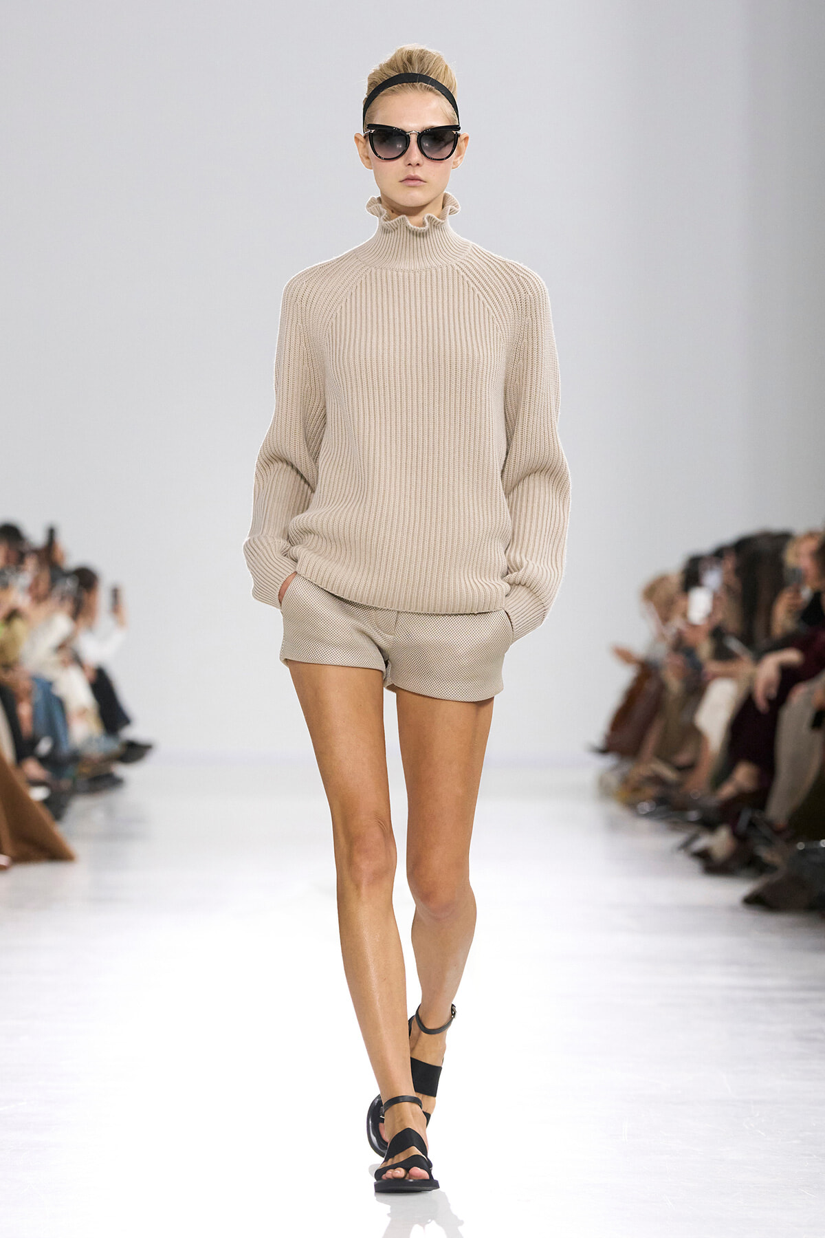 Model walking a runway in a beige ribbed turtleneck sweater and matching shorts, with black sunglasses and a headband.
