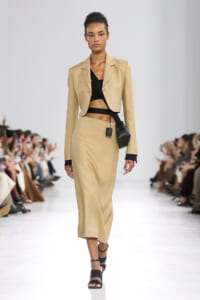 Female model walks a runway wearing a tan tailored blazer and matching midi skirt with a black cropped top and a small black crossbody bag.