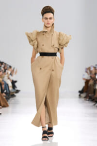 Model on a runway wearing a beige trench-style dress with oversized ruffled sleeves and a black waist belt.