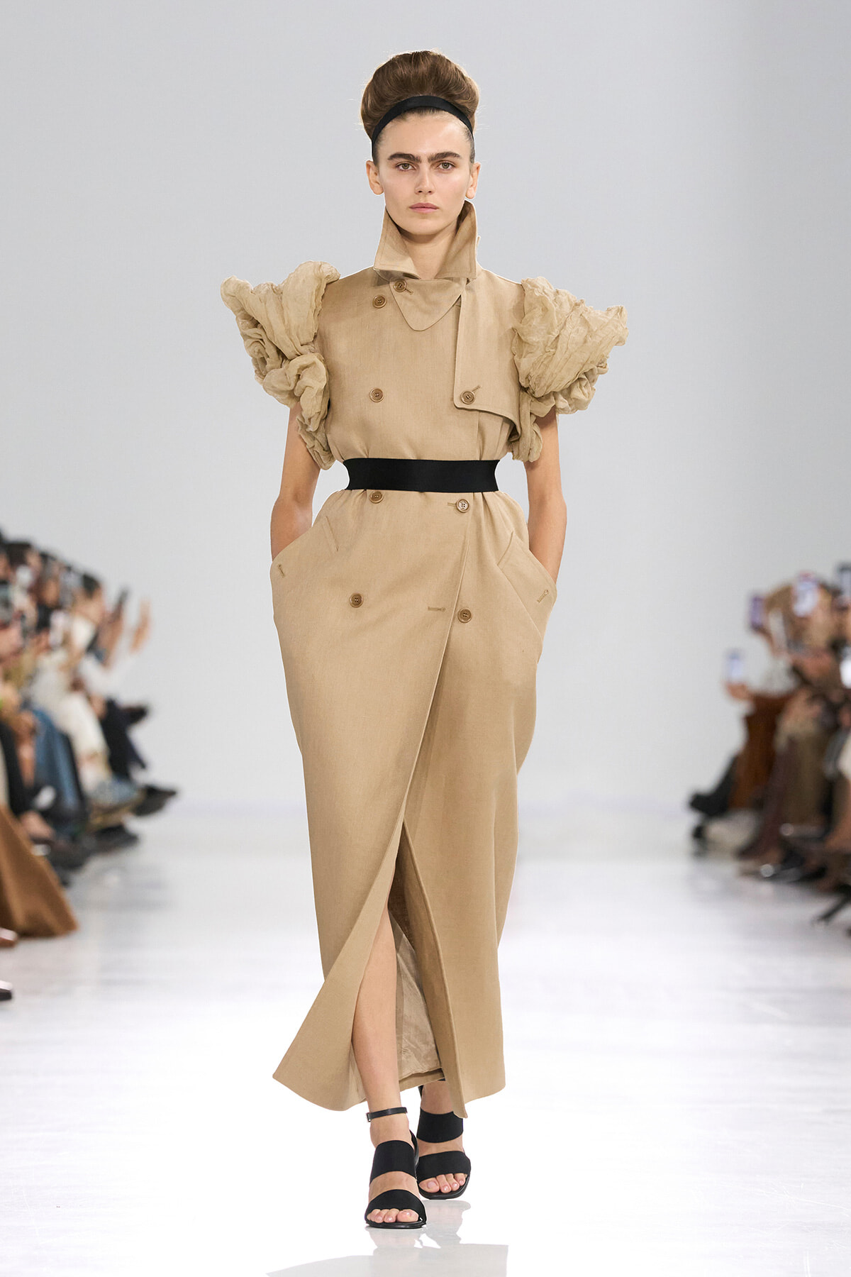 Model on a runway wearing a beige trench-style dress with oversized ruffled sleeves and a black waist belt.