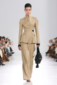 Female model walks the runway in a beige tailored suit with a structured, peplum-style blazer and matching wide-leg pants, carrying a black handbag.