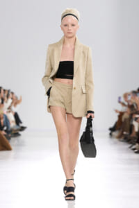 Model walking on a runway in a beige tailored blazer with matching shorts, a black bandeau top, and black sandals, carrying a small black bag.