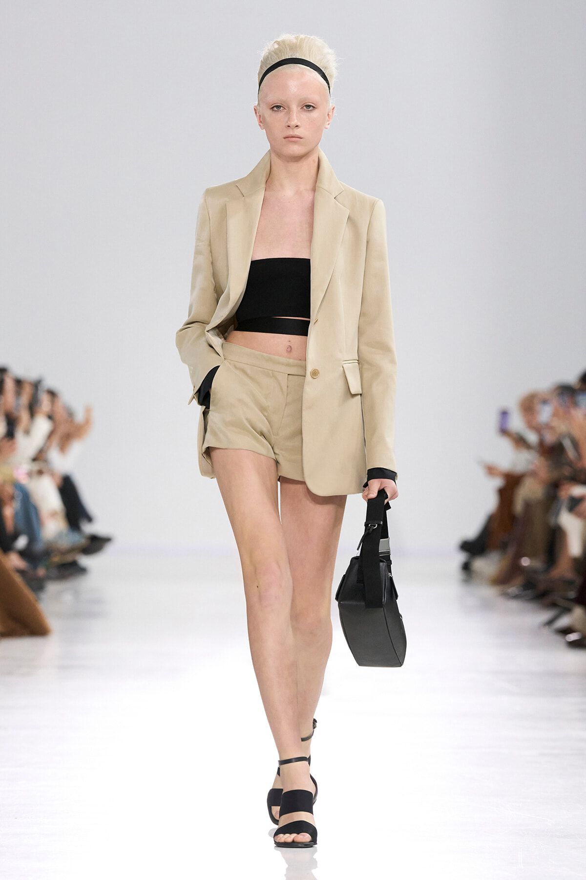 Model walking on a runway in a beige tailored blazer with matching shorts, a black bandeau top, and black sandals, carrying a small black bag.