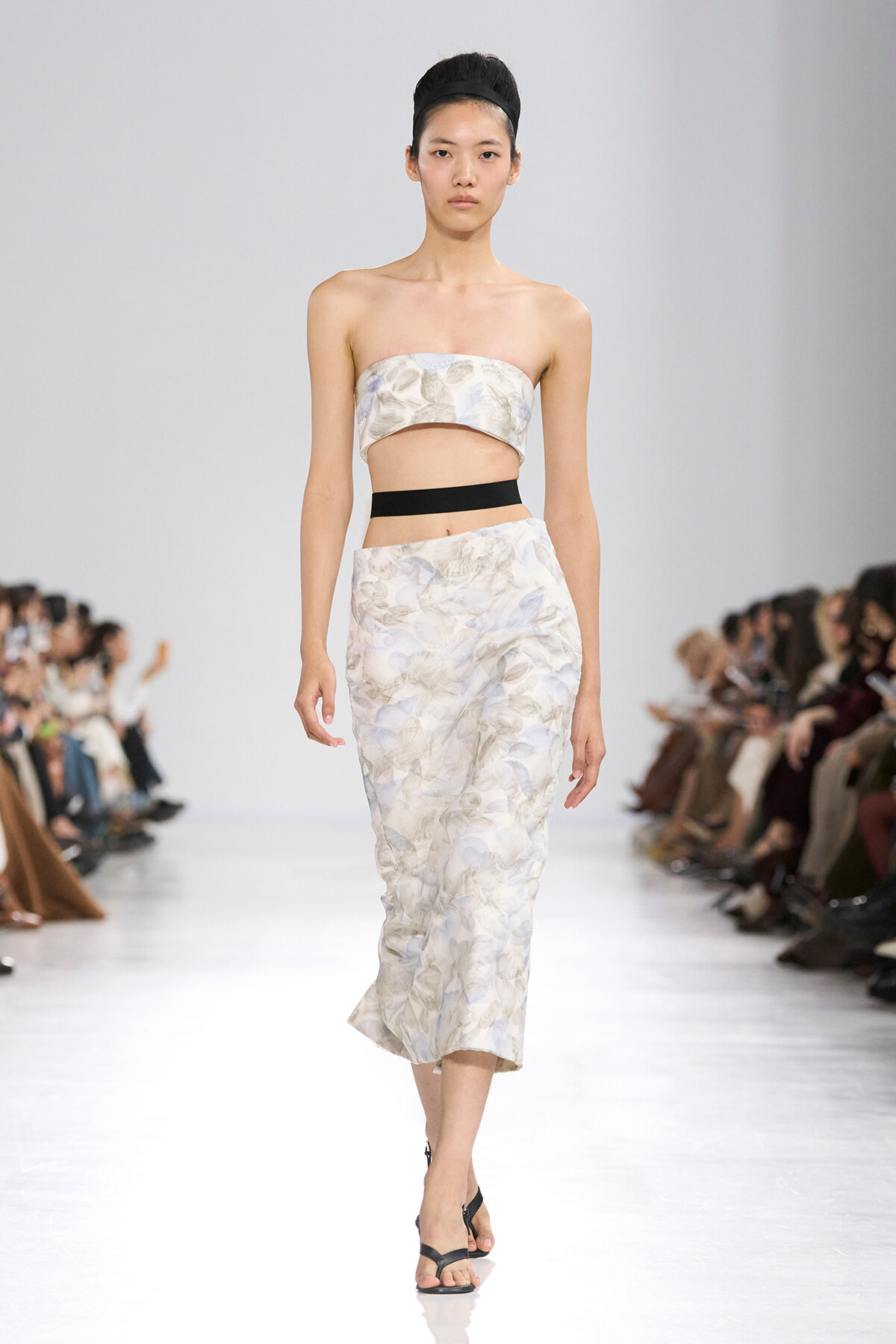 Model walks the runway wearing a strapless floral bandeau top with a matching midi skirt and a black waist band, black sandals, and a headband.
