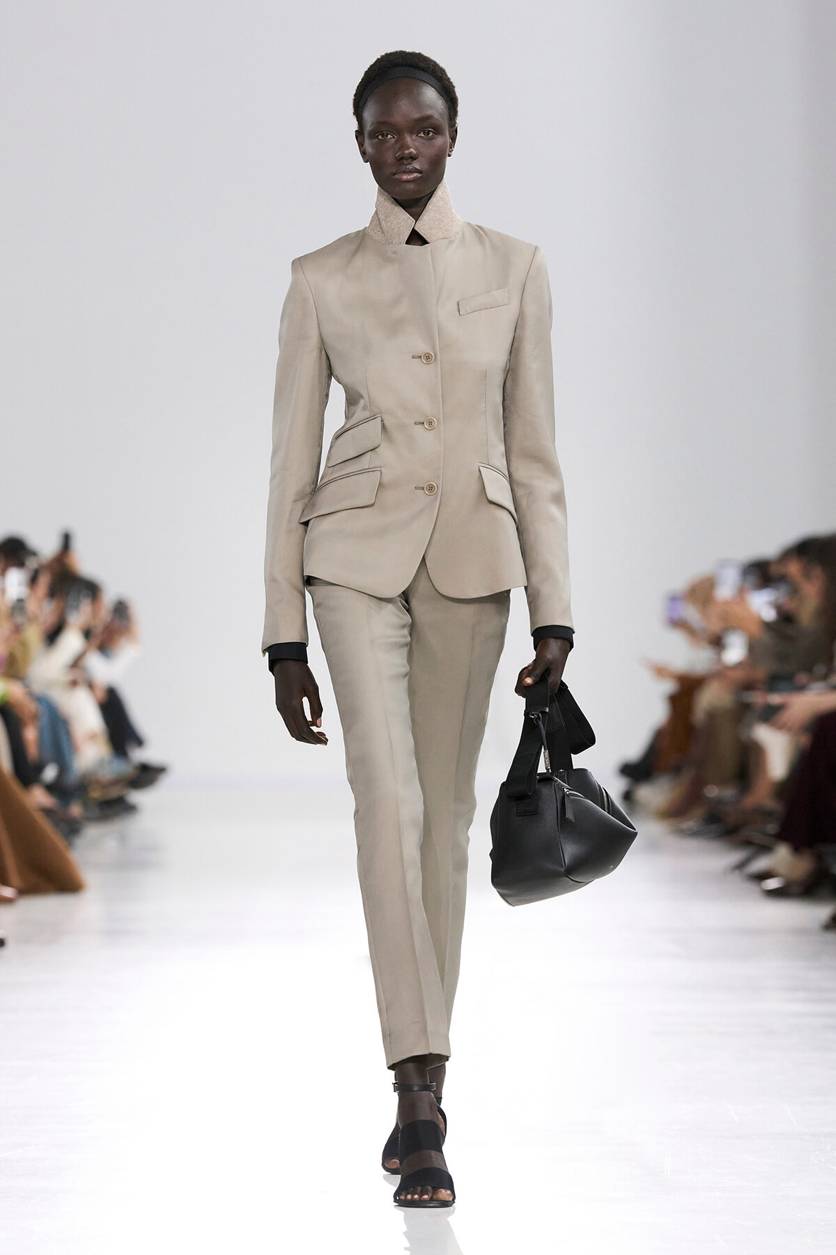 Female model wearing a beige tailored suit with a high collar, walking a runway with a black handbag.