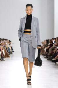 Model on a runway wearing a light gray suit with a black cropped turtleneck, carrying a black handbag.