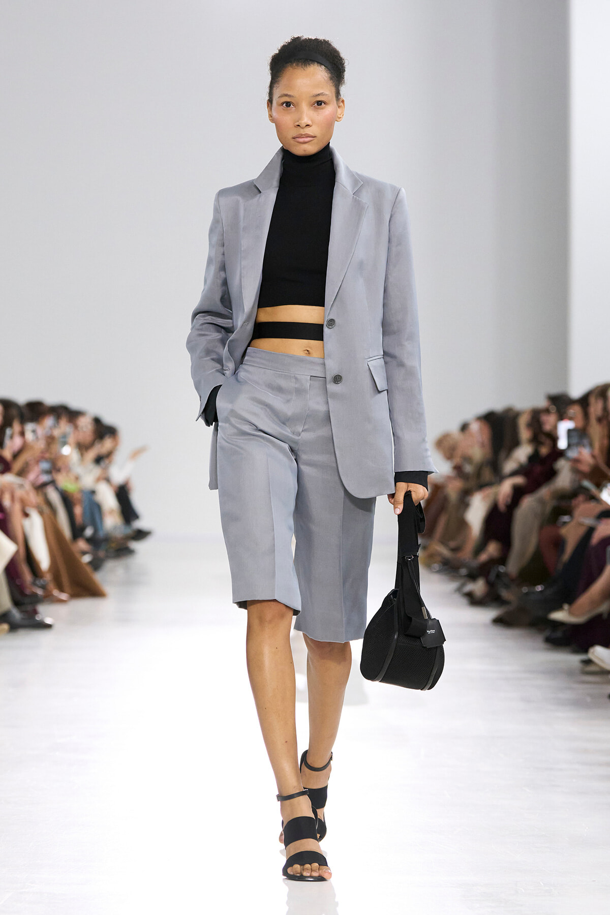 Model on a runway wearing a light gray suit with a black cropped turtleneck, carrying a black handbag.