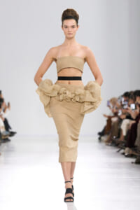 Model on a runway wearing a tan strapless bandeau top with a high-waisted beige pencil skirt featuring large ruffled fabric at the hips, black waist band, and black sandals.