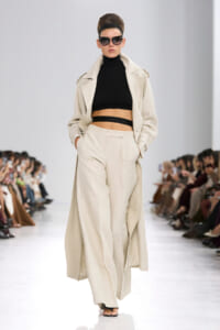 Model on a runway wearing a beige, long trench coat over a black cropped top and matching wide-leg trousers, with sunglasses and an updo.