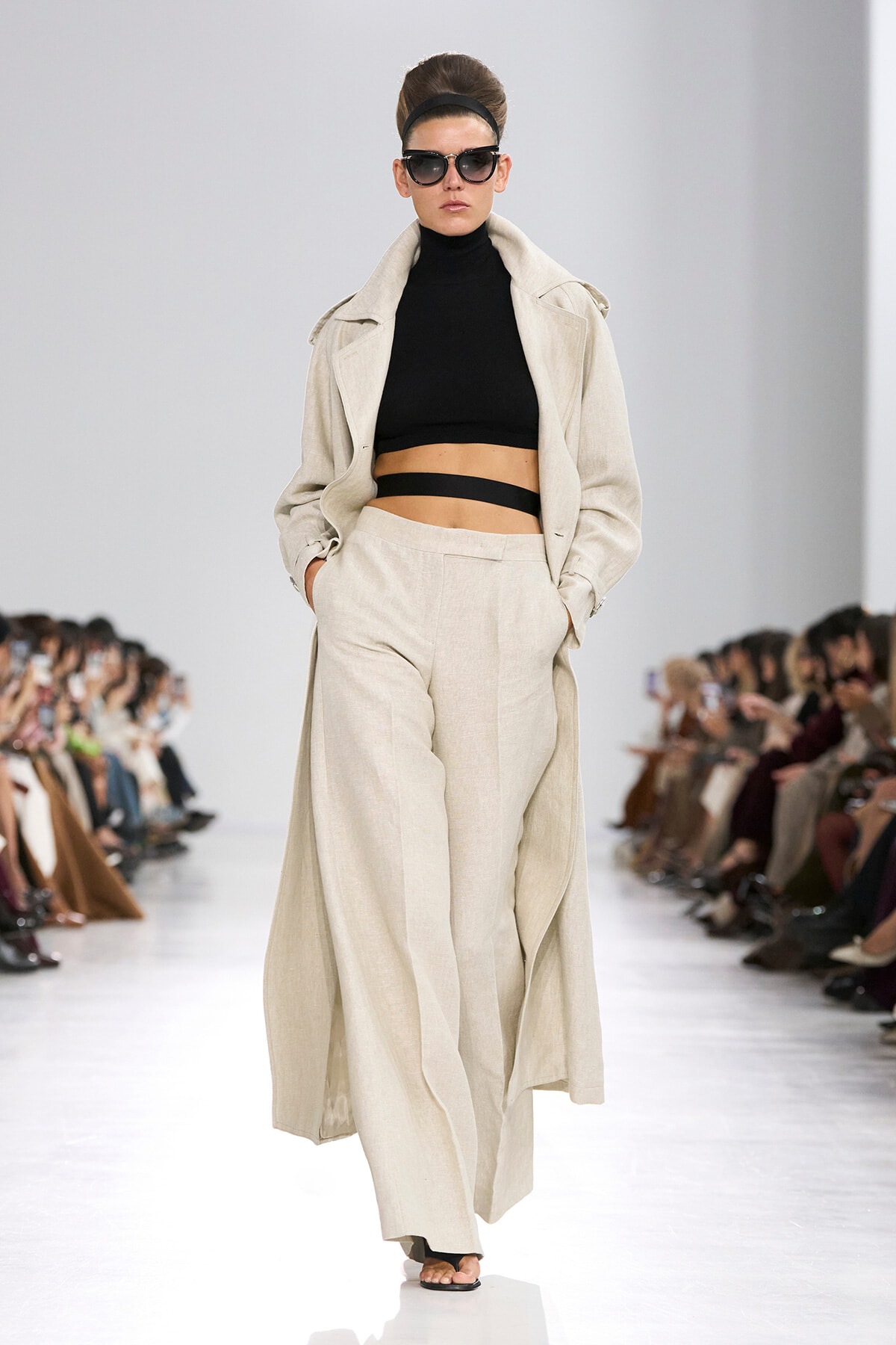 Model on a runway wearing a beige, long trench coat over a black cropped top and matching wide-leg trousers, with sunglasses and an updo.