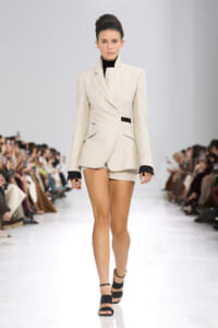 Female model walks a fashion runway wearing a beige tailored blazer styled as a short dress, with black turtleneck and black heeled sandals.
