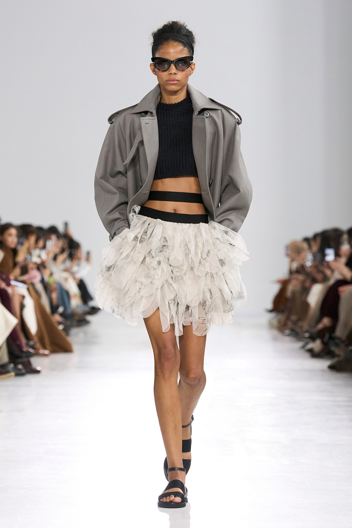 Model walking runway in a gray oversized trench, black cropped sweater, and a voluminous white feathered skirt with black belt, wearing black sunglasses.