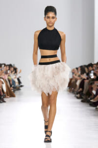 Model walking a fashion runway in a black knit halter crop top and a pale pink layered tulle skirt with black waistbands; audience seated along the sides.