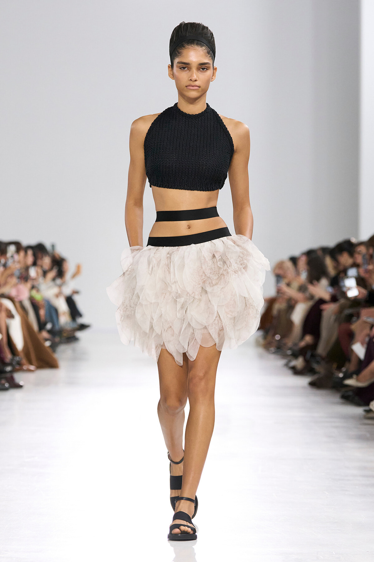 Model walking a fashion runway in a black knit halter crop top and a pale pink layered tulle skirt with black waistbands; audience seated along the sides.
