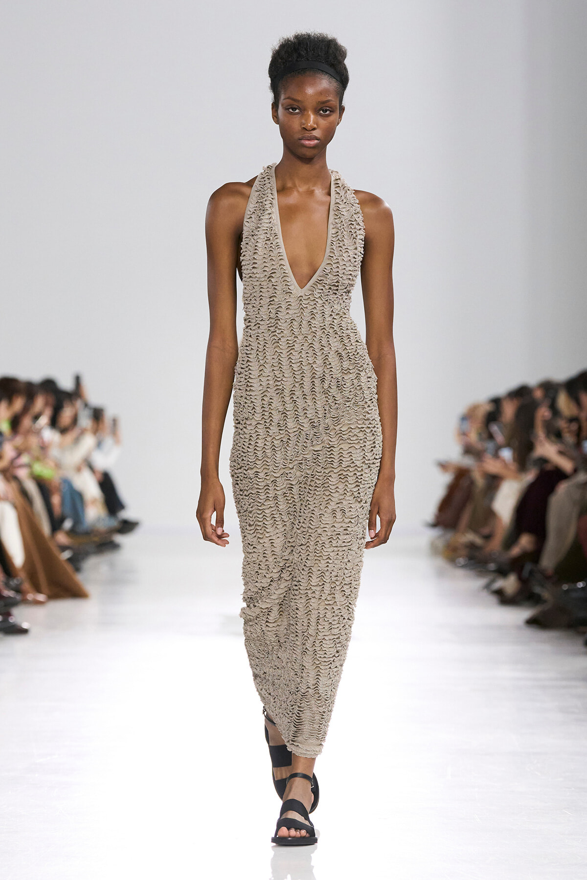 Model walks a fashion show wearing a beige textured halter-neck dress with deep V and black sandals; audience seated along the runway in the background.