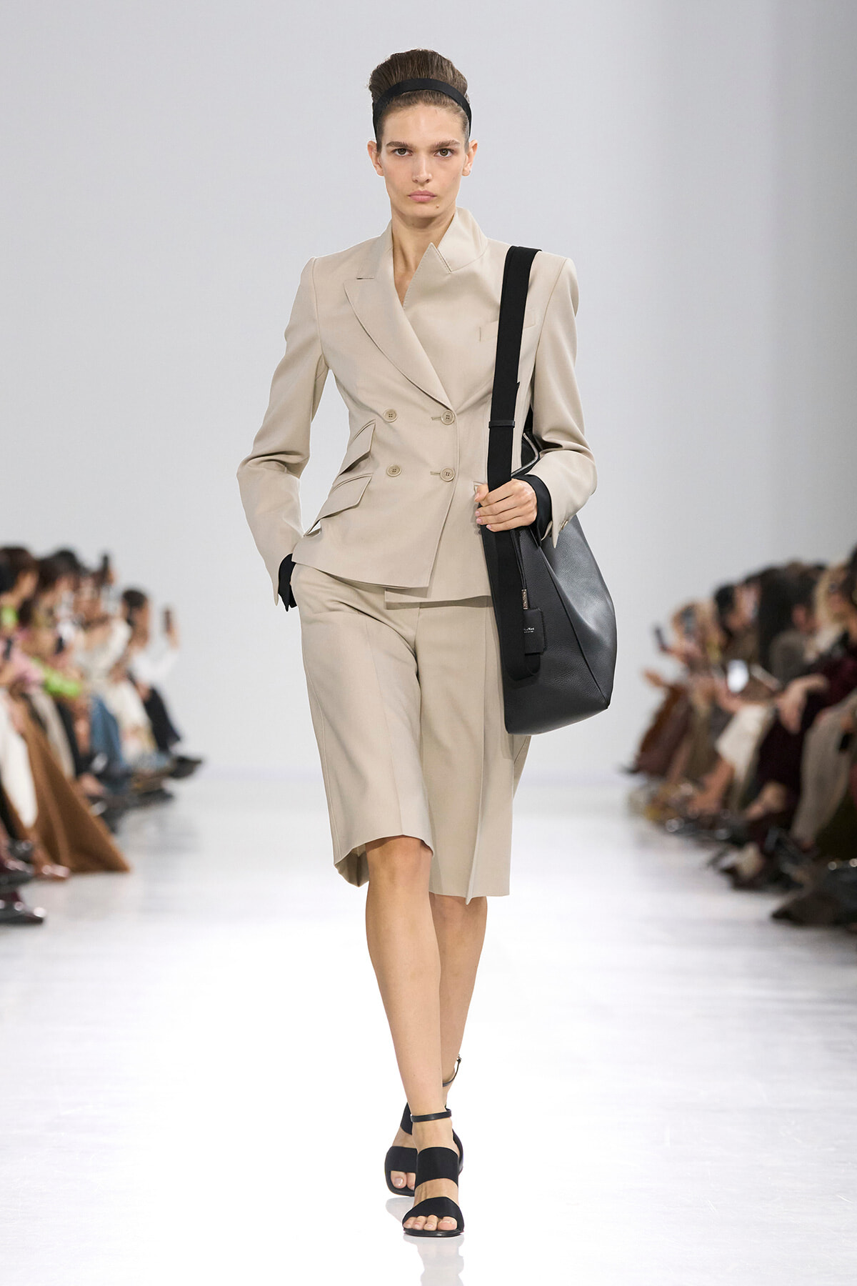 Female model on a runway wearing a beige double-breasted blazer and matching knee-length shorts, with a black headband and large black shoulder bag.