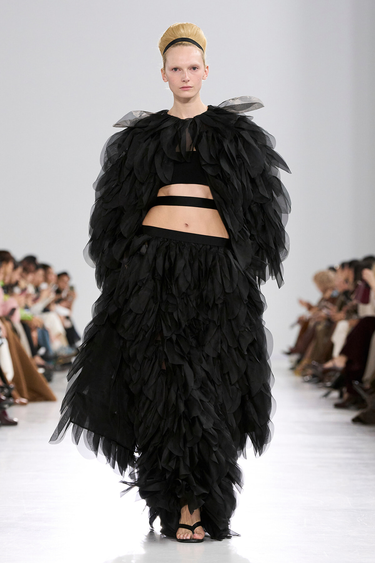 Model on a runway wearing a voluminous black feathered gown with a cropped top and waist belt, hair in a headband.