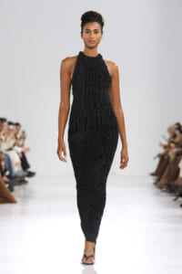 Model walking runway in a fitted black textured halter-neck dress with black sandals against a white backdrop.