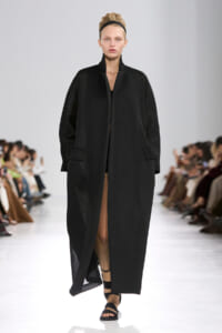 Model on a runway wearing a long black textured coat with a high collar and front pockets, paired with black strappy sandals.