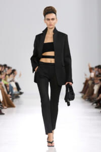 Model walking a runway in a black textured suit with a cropped black bandeau top and cutout straps, carrying a small black handbag.